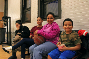 Martinez sitting between her two children, age 14 and 9, with a basketball in her lap. Martinez's daughter and her friend (far left) left the game and returned with a frozen yogurt to share.