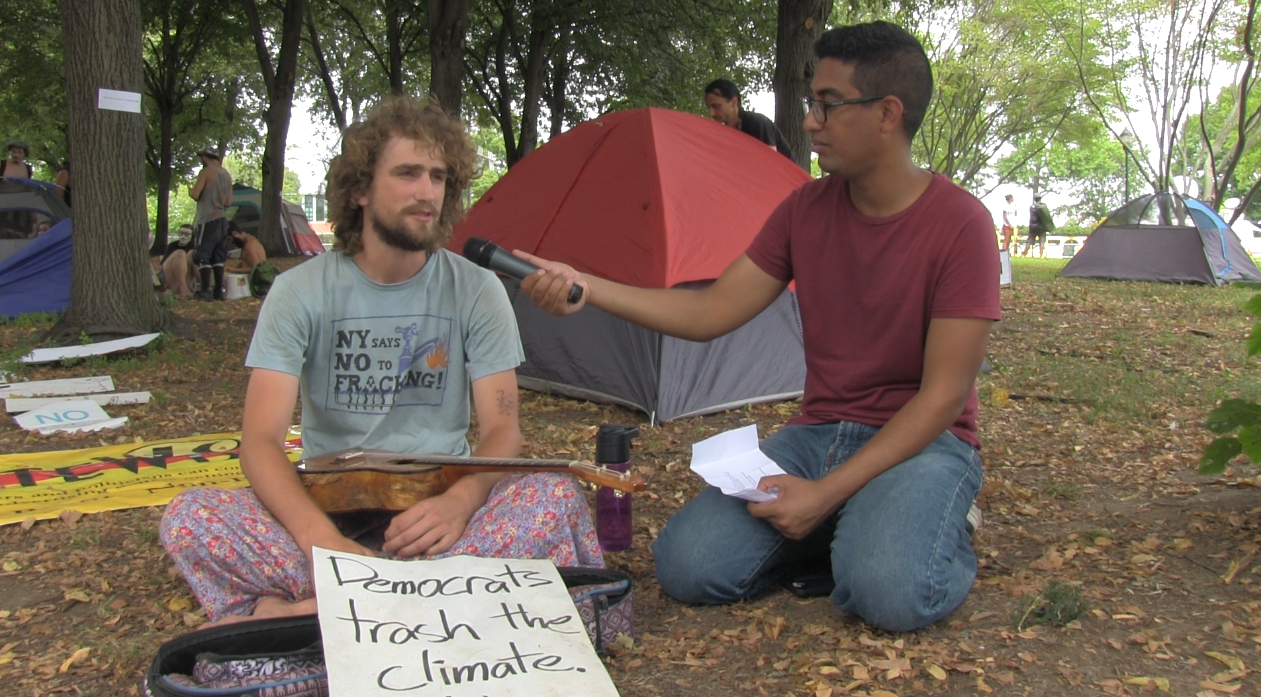 Tom Moore, a Bernie Sanders supporter, speaks with Abdelfattah. He has a sign in front of him that says "Democrats trash the climate." (Photo by Jenny Montasir, GroundTruth)