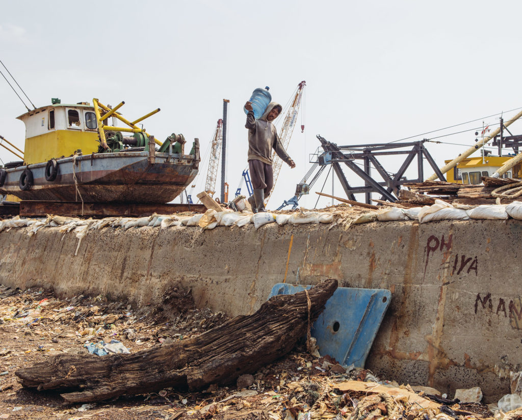 A worker in Pluit district of North Jakarta carries a gallon of drinking water atop the seawall that flanks the area from the sea. Currently, this area is already below the sea level. (Photo by Muhammad Fadli/GroundTruth)