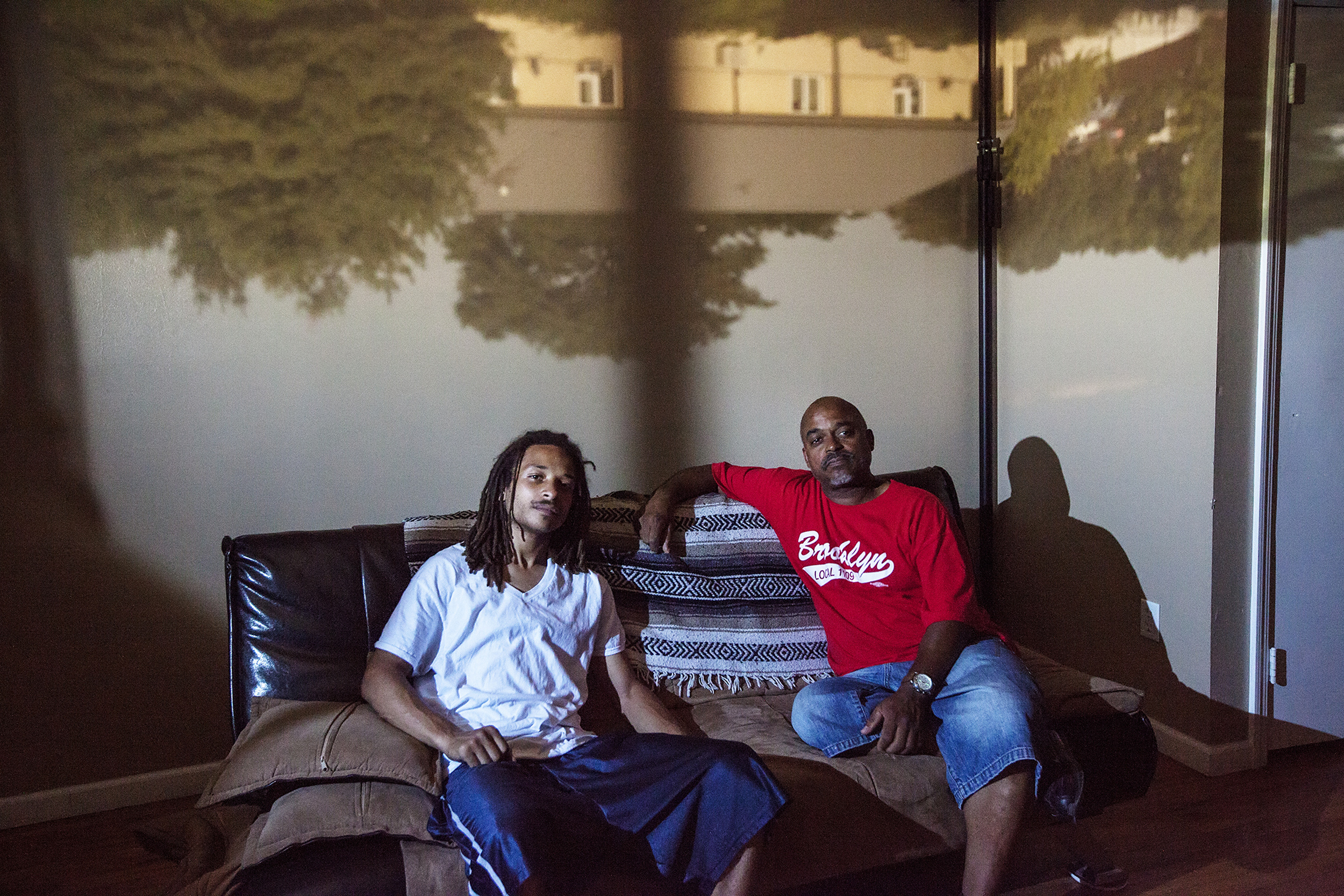 Michael Cathlin, 26, poses for a portrait with his step father, Michael Burke-Andrade, in East New York, NY on July 25, 2015. "Thankfully I walked in [the delivery room] right before my son started crying. So I was there for the whole thing but it was the fact that the mother didn't want me there." Raising East New York (RENY) is a portraiture project about Black fathers where the photographer, Phyllis B. Dooney, uses a photographic technique called Camera Obscura to project the outside streets into the subjects apartment in real time.