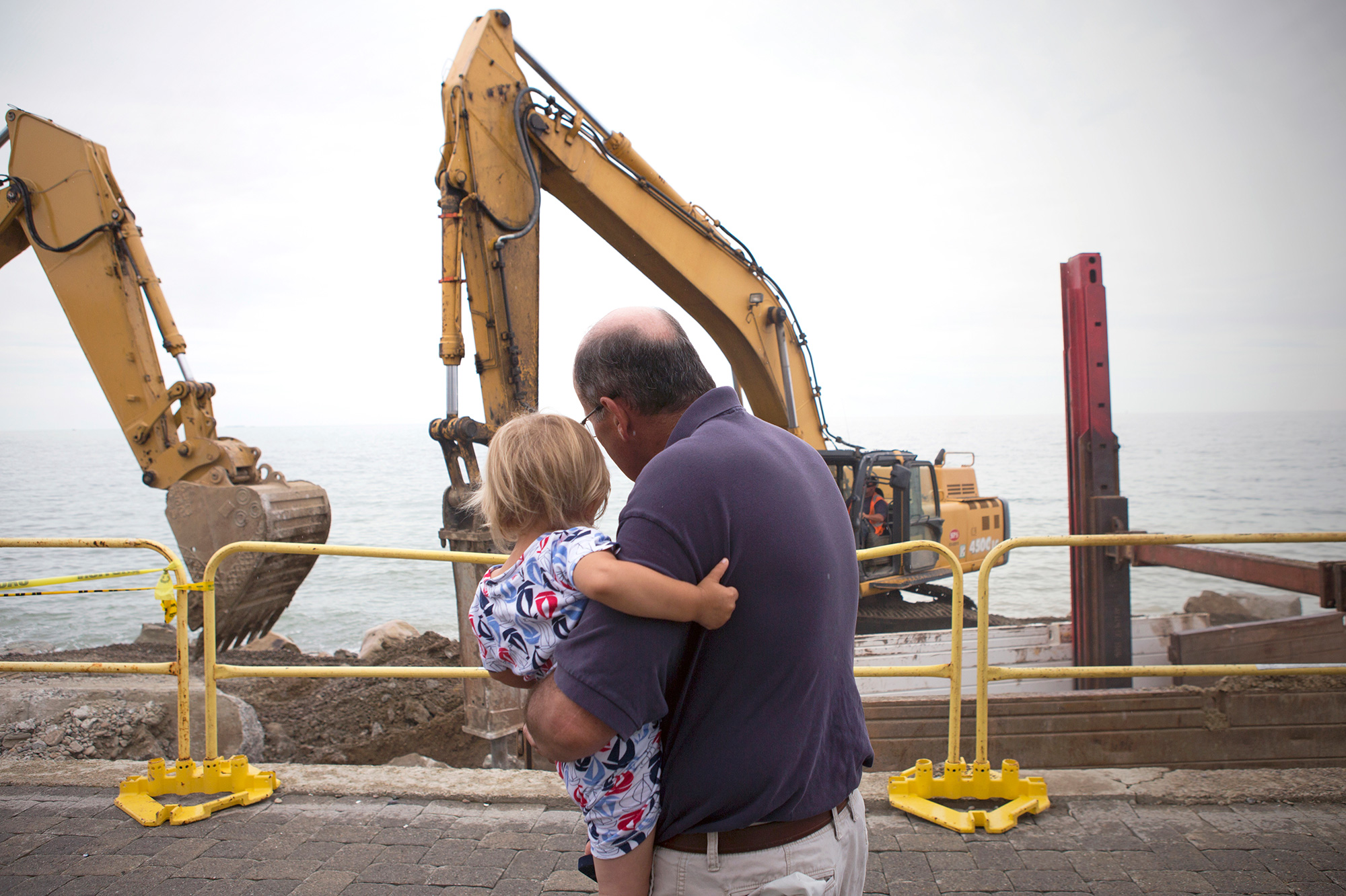 Scituate resident Peter Caruso observes the construction of a new seawall in Scituate with his grandson, Santo Arias. The town hopes that bolstering the seawall will mitigate the severity of winter storms and the effects of sea level rise. “It is a tough spot to try and live and the storms are more frequent than they use to be,” says Caruso. (Photo by Lauren Owens Lambert/GroundTruth)