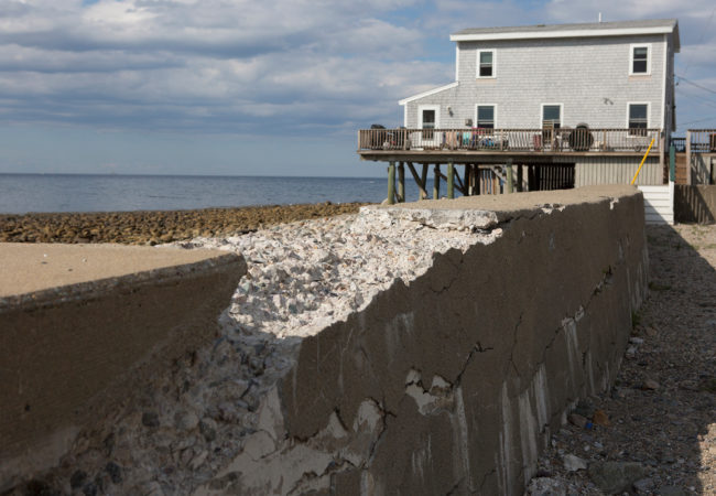 Next to a raised home in Scituate, a seawall crumbles—the result of years of ocean waves pummeling the shores. Most coastal towns along the Massachusetts South Shore have man-made sea walls in place to help protect the ocean front from storm surges. But recent storms have breached the seawalls and weakened them for future storms. (Lauren Owens Lambert/GroundTruth)