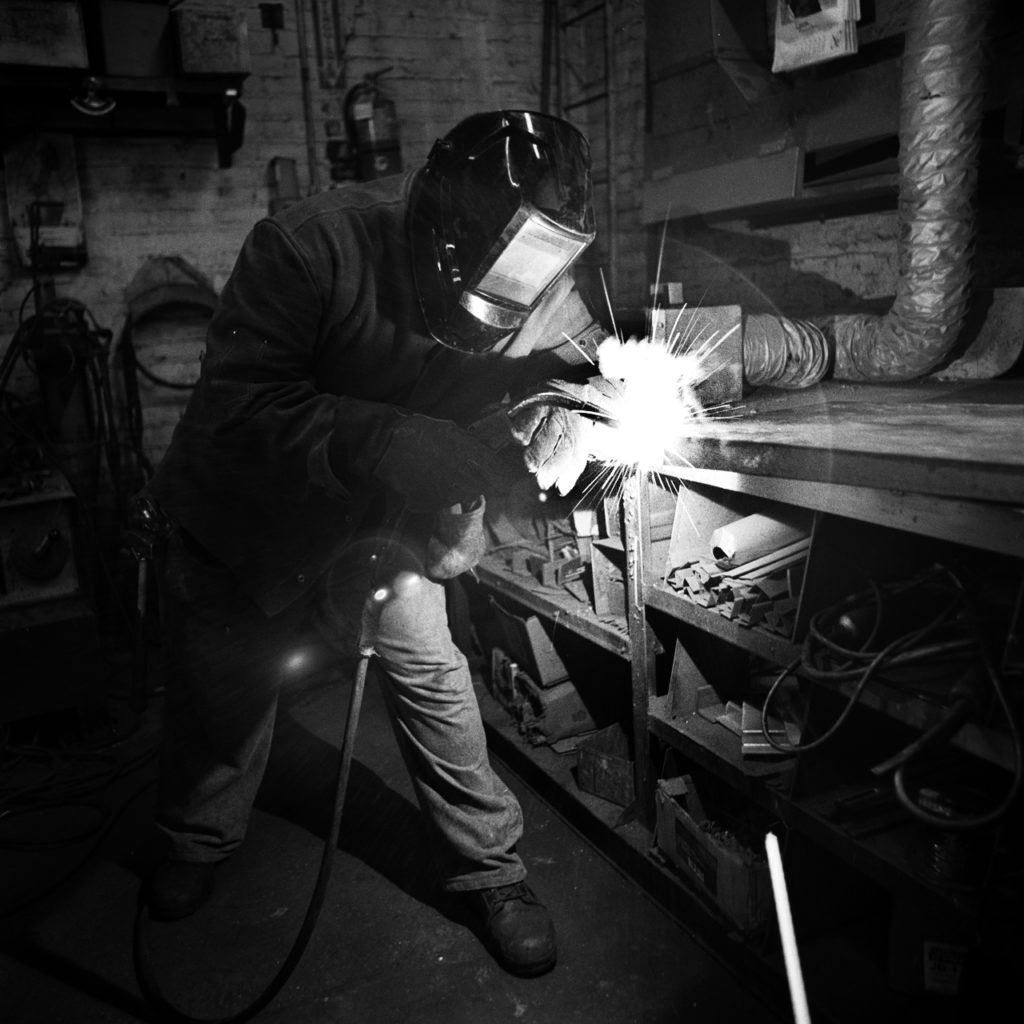 Derrick Green, from Gentlemen Sheet Metal, works at his welding station. A former IT worker, Derrick remembered the huge annual packets he had to fill out to prove to management his case for a raise. Today, he enjoys sitting back and letting his union, Local 28, fight for his prevailing wage. A makeshift memorial for Thomas R. Dolphin was put up near the intersection of Meeker Avenue and Gardner Avenue in Greenpoint, Brooklyn, N.Y. (Photo by Dakota Santiago/GroundTruth)