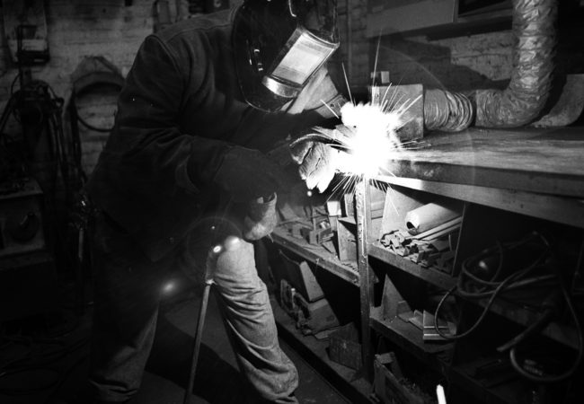Derrick Green, from Gentlemen Sheet Metal, works at his welding station. A former IT worker, Derrick remembered the huge annual packets he had to fill out to prove to management his case for a raise. Today, he enjoys sitting back and letting his union, Local 28, fight for his prevailing wage. A makeshift memorial for Thomas R. Dolphin was put up near the intersection of Meeker Avenue and Gardner Avenue in Greenpoint, Brooklyn, N.Y. (Photo by Dakota Santiago/GroundTruth)