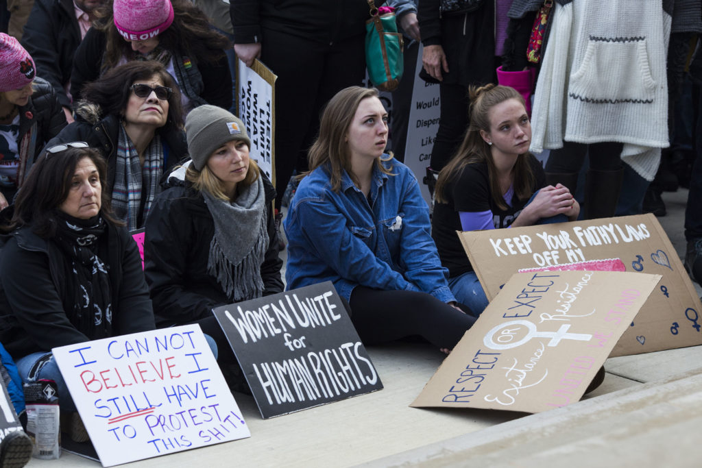 Young women sit along the steps outside of the Michigan State Capitol as a representative of Planned Parenthood speaks about the possible funding losses under the new presidential administration, during the Women's March on Lansing in Lansing, Mich., on Saturday, January 21, 2017. (Photo by Brittany Greeson/GroundTruth)
