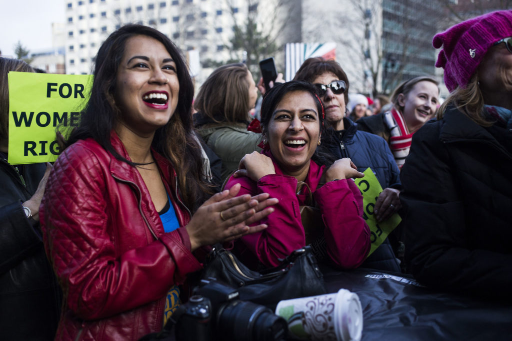 Sharmin Begum, 23, right, and Tahrima Khanom, 26, left, both of Hamtramck, Mich., laugh at a speaker's remarks during the Women's March outside of the Michigan State Capitol Building in Lansing, Mich., on Saturday, January 21, 2017. (Photo by Brittany Greeson/GroundTruth)