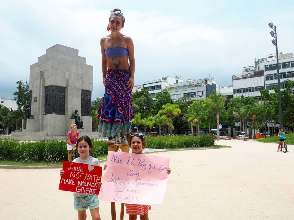 A marcher on stilts and two children with their signs pose for a photo. The sign on the right reads, "There is no peace that lasts without justice and equality for all." (Photo by Catherine Osborn/GroundTruth)
