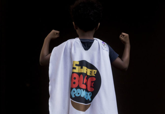 Matias Melquiades, 5, poses with the "super black power" cape at the Black Power Girls Annual Fair. This and other representative toys and dolls are made by the company created by his parents, Leandro and Jaciana. (Photo by Leonardo Coelho/GroundTruth)