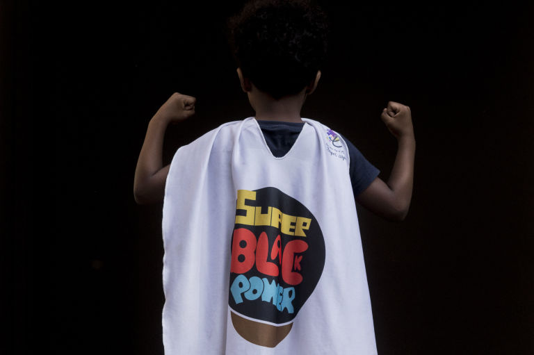 Matias Melquiades, 5, poses with the "super black power" cape at the Black Power Girls Annual Fair. This and other representative toys and dolls are made by the company created by his parents, Leandro and Jaciana. (Photo by Leonardo Coelho/GroundTruth)