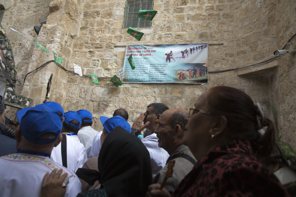 Egyptian Christian worshippers stand on the rooftop of the The Church of the Holy Sepulchre during Good Friday as they pass by a poster showing images from 2015 released by the Islamic State of Iraq and the Levant (ISIL) – Christian migrant workers that they had kidnapped in the city of Sirte, Libya (Photo by Heidi Levine)