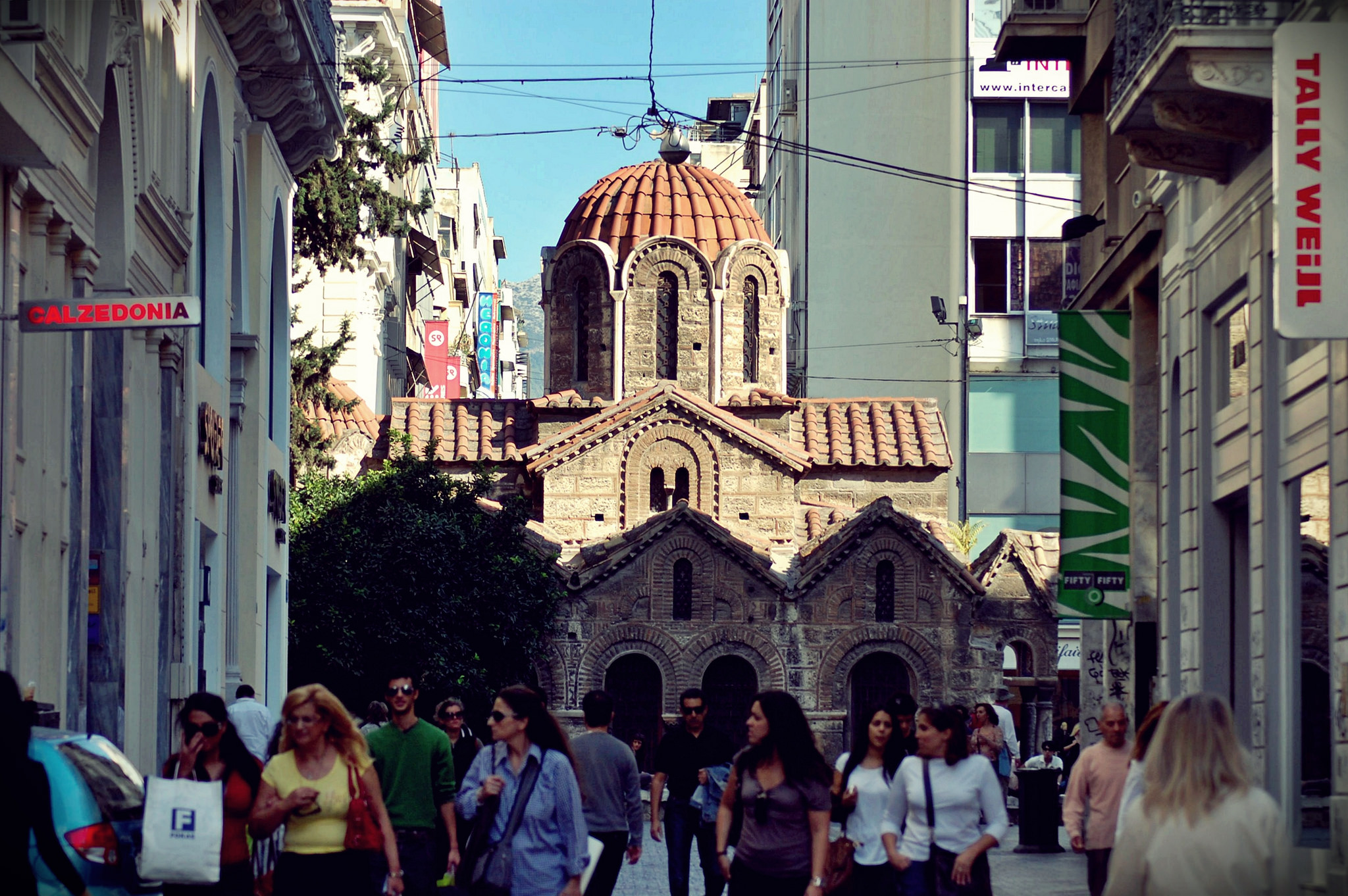 People shop on a busy street with Athen's 11th-century Byzantine Church of Panaghia Kapnikarea behind them. (Photo by Ananabanana/Flickr User)