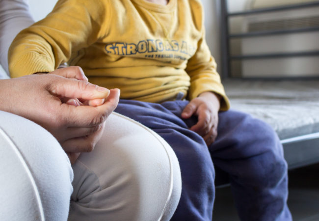 In the Sindos apartment complex, Alia Habesh, 25, holds her youngest son’s hand while sharing her story of their journey from Aleppo, Syria to Thessaloniki, Greece. Habesh fled Aleppo in 2012, leaving behind a life as a wedding gown embroiderer. (Photo by Catherine Clark/GroundTruth)