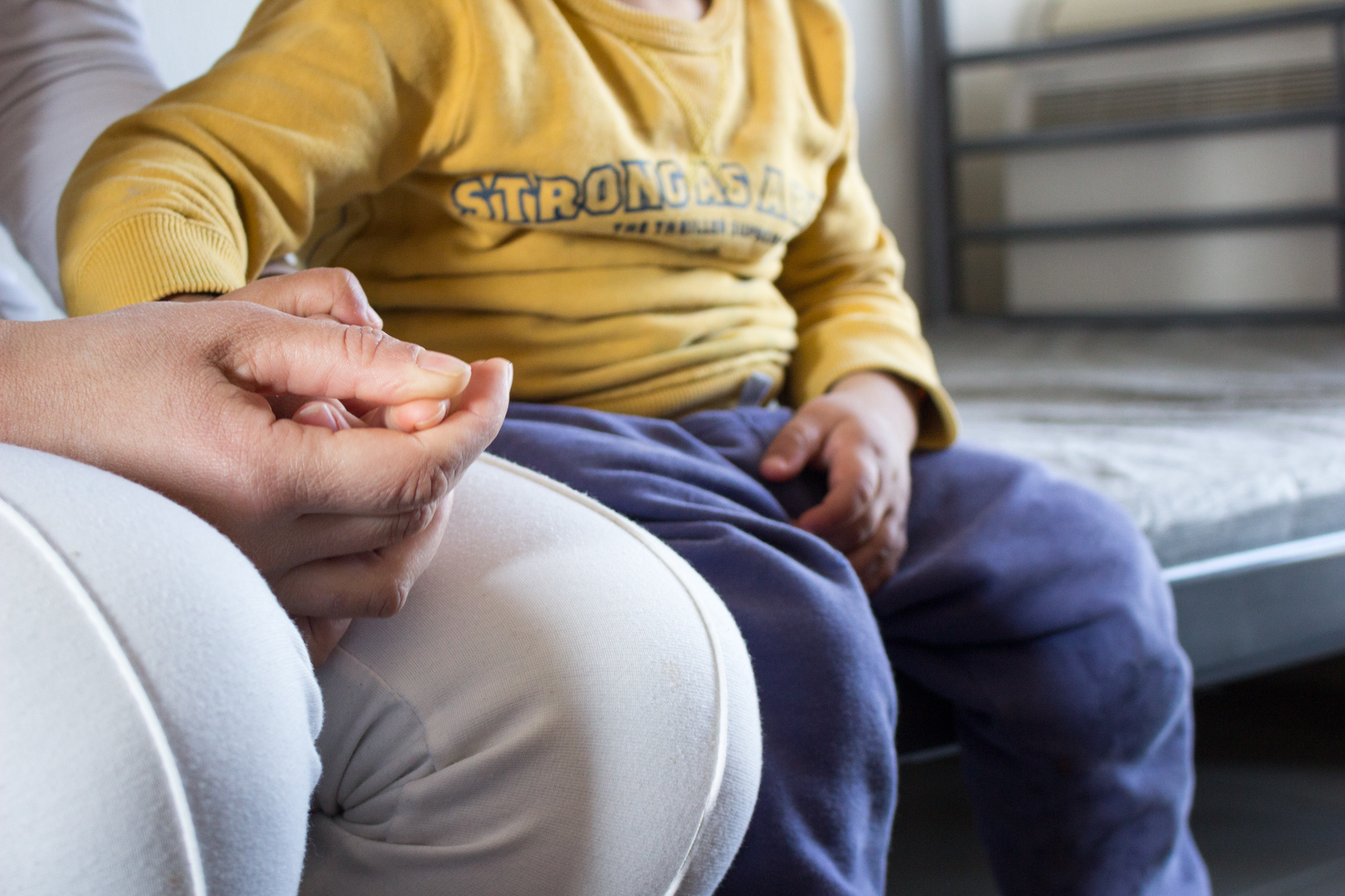 In the Sindos apartment complex, Alia Habesh, 25, holds her youngest son’s hand while sharing her story of their journey from Aleppo, Syria to Thessaloniki, Greece. Habesh fled Aleppo in 2012, leaving behind a life as a wedding gown embroiderer. (Photo by Catherine Clark/GroundTruth)