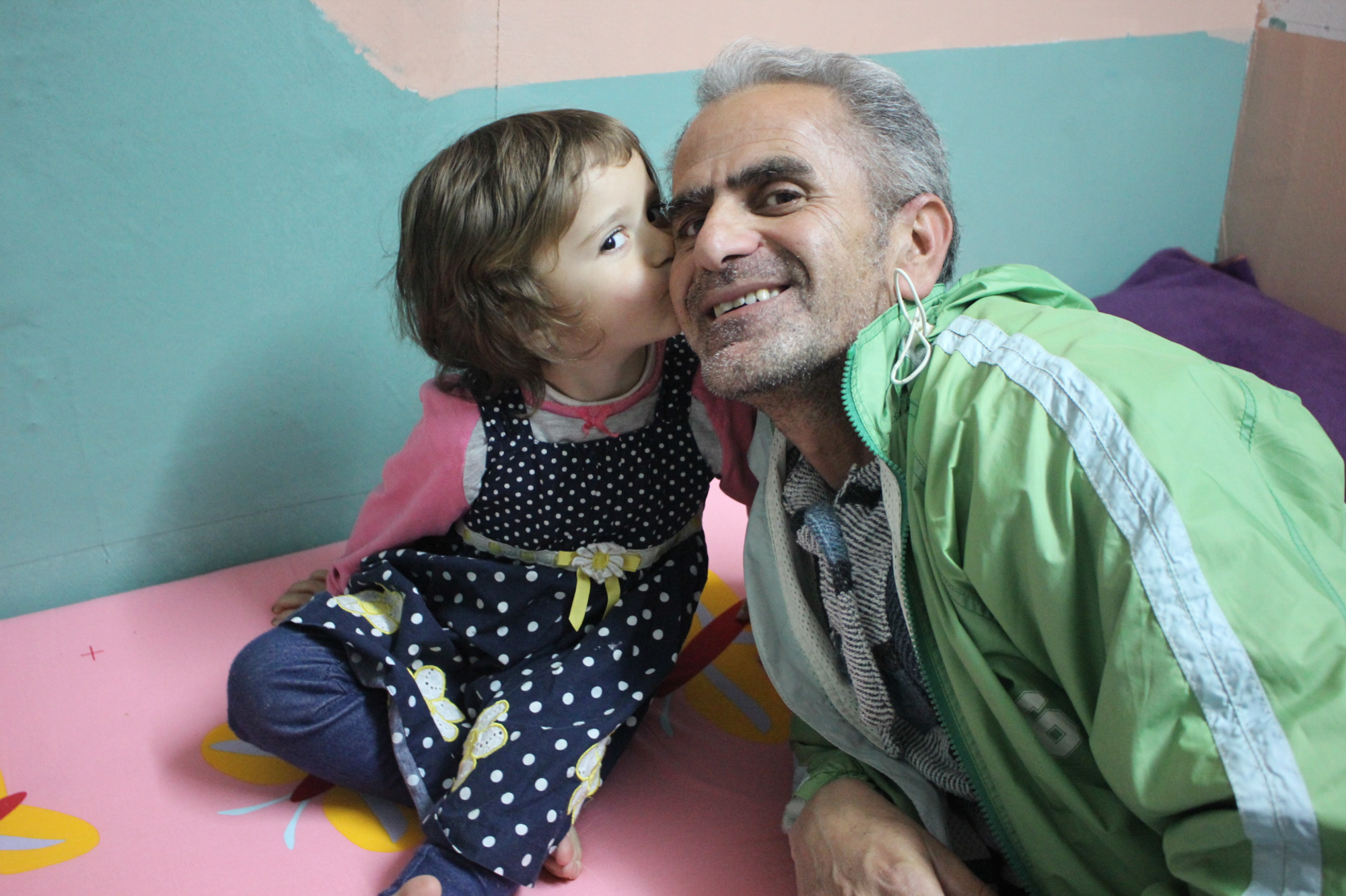 A young Syrian girl kisses her father’s cheek inside their colorfully decorated room at Oinofyta, a refugee site outside of Athens, Greece. (Photo by Alexandra Meeks/GroundTruth)