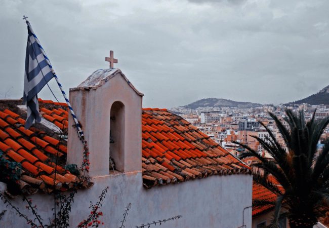 A Greek flag hangs from the roof of a small church overlooking Athens. (Photo by Mstyslav Chernov/Wikimedia Commons)