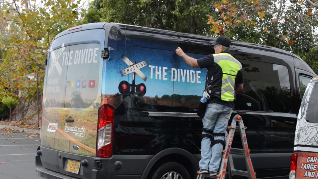 JC strips the "Crossing the Divide" wrap off the van. (Screenshot)