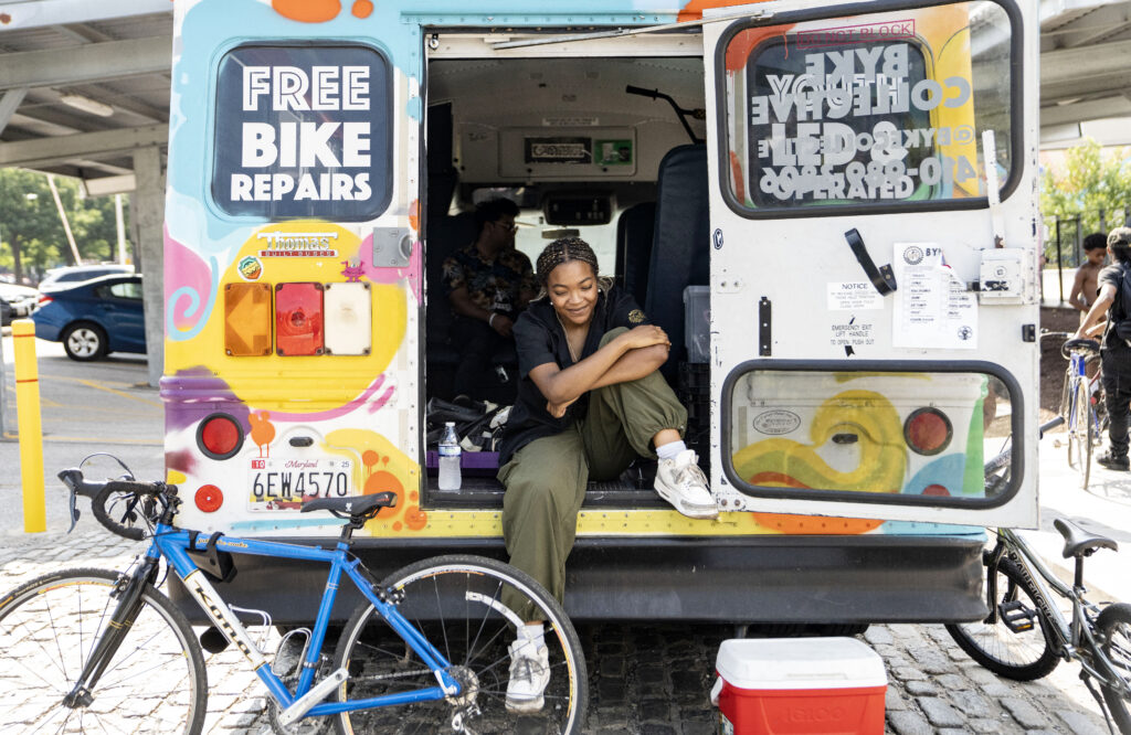 A young woman wearing army green pants and white sneakers smiles quietly while sitting on the edge of a colorful bus. There's a bike and a cooler in front of her.