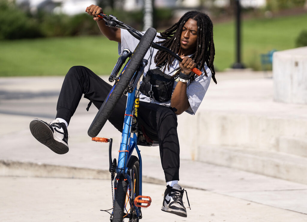 A young man with brown skin and long braids pops a wheelie on a blue bicycle.