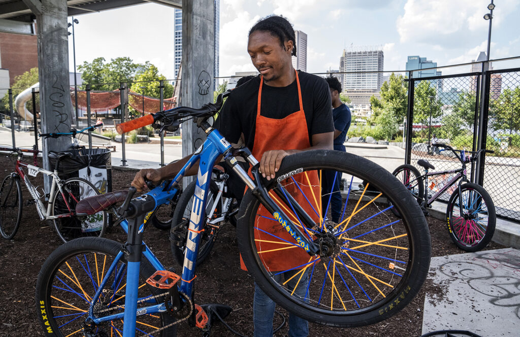 A young man with brown skin wearing an orange apron repairs a bicycle. There are many bikes around him.