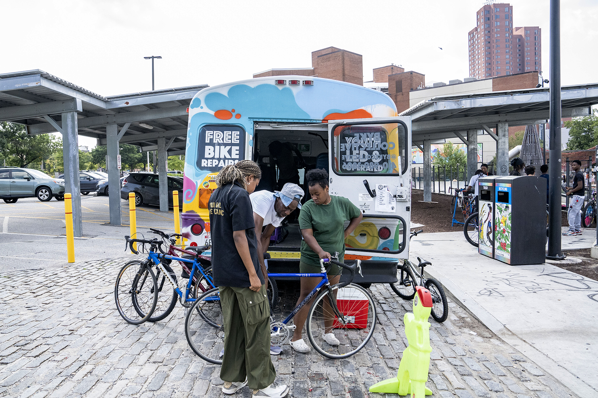 Three mechanics (one male, two female) repair a bicycle for a resident in front of BYKE's mobile bus in the Inner Harbor.