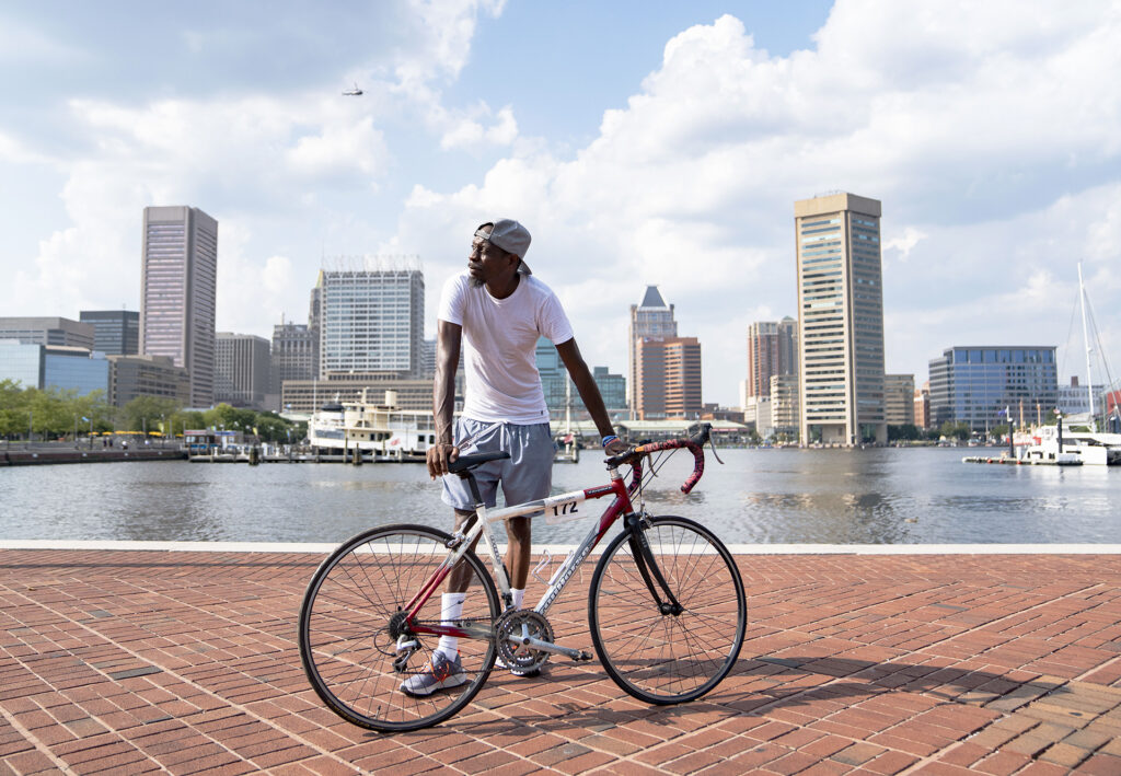 A man with brown skin wearing a backwards baseball cap, a white tee shirt, shorts and sneakers with socks leans on his bicycle in Baltimore's Inner Harbor.