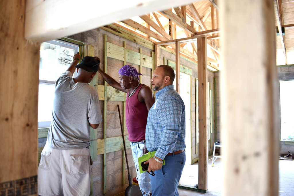 Two construction workers install a window in a new house while a man in a button-down shirt and blue trousers watches.