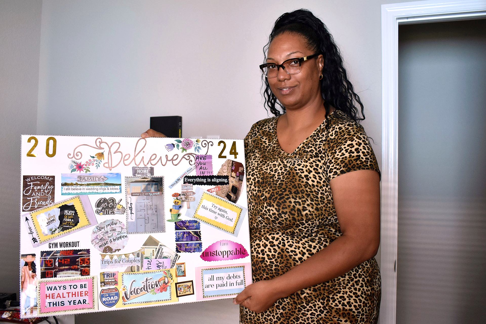 A woman with brown skin, glasses and a long black ponytail holds up her 2024 vision board.