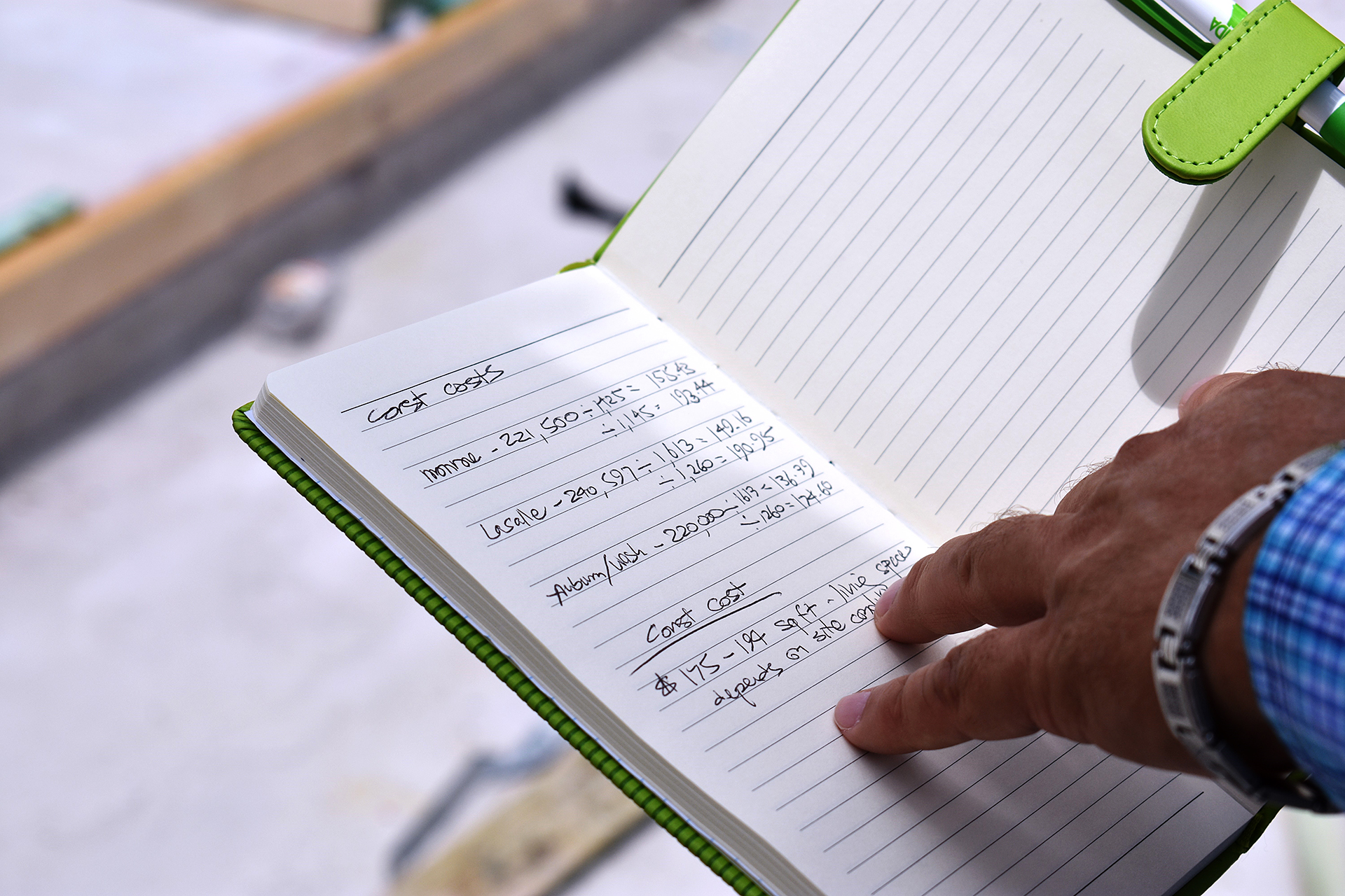 A notebook is open showing building costs and there is a man's hand pointing at the numbers.