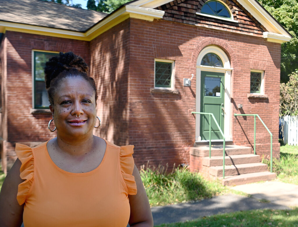 A woman with brown skin, freckles, a nose ring and earrings, who is wearing an orange shirt stands in front of a brick house.