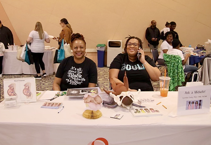 This is a photo of two women with brown skin, both wearing black tee shirts, sitting at a table ready to speak to prospective and new parents.