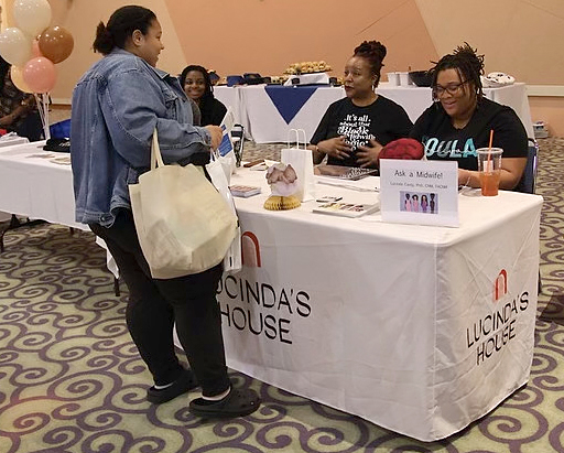 Two women with brown skin and wearing black tee shirts sit a table with "Lucinda's House" branding and speak with a woman standing across from them.