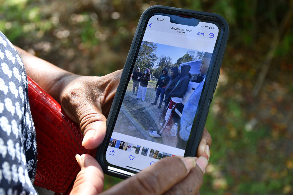 A woman's hands hold a cell phone with an image of young men, most wearing hoodies, listening to someone speak on the farm.