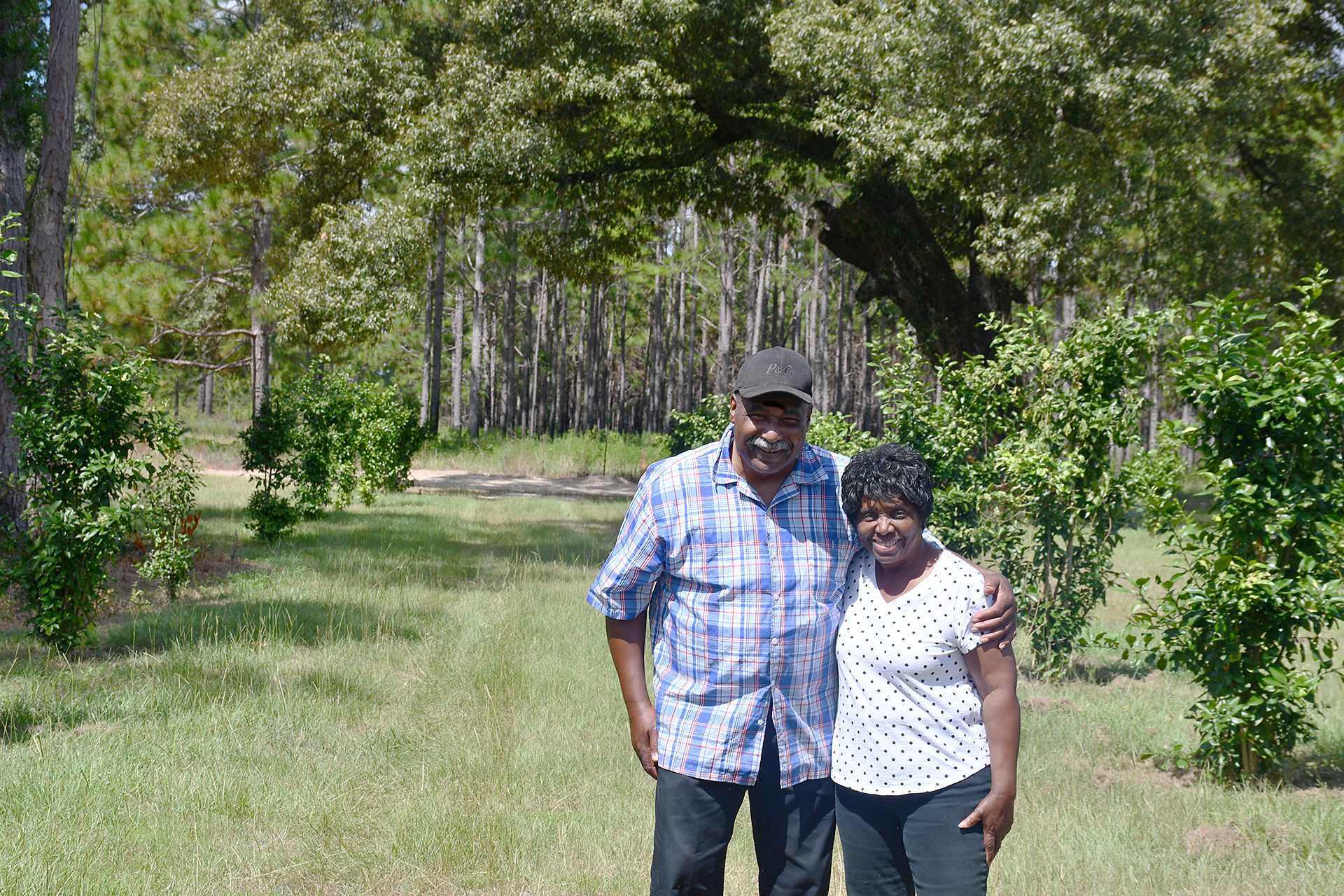 A man wearing a hat and his wife, both with brown skin, stand with their arms around each other on their farm.