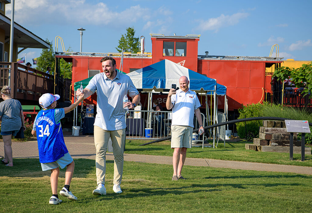 A white man in a light blue shirt with an expressive face dances with a young boy wearing a basketball jersey while another man takes a photo.