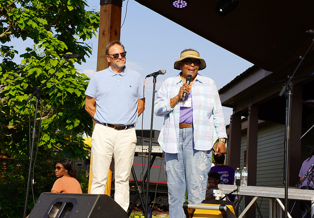 A man with pale skin wearing a polo shirt, khakis and sunglasses stands to the left of a woman wearing jeans, a purple tee shirt and an open button down. She's wearing a sunhat and speaking into a microphone.